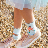A child wearing pink trainers and a blue dress/skirt is wearing the light blue pair of Frugi Frill Socks 2 Pack- Enchanted Forest, with the ice skating fairy print