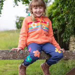 A child sat on a wooden fence, wearing the Frugi Perrin Parsnip Jogger - Navy Blue/Flowers, and a Frugi hoody