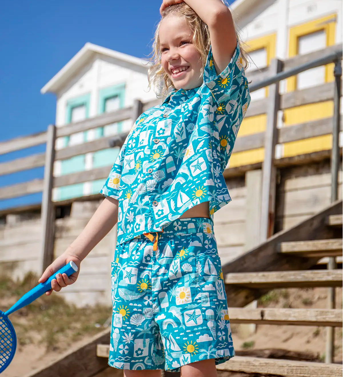 Child wearing Frugi's Kai shorts in blue with repeating beach inspired designs with beach huts in the background