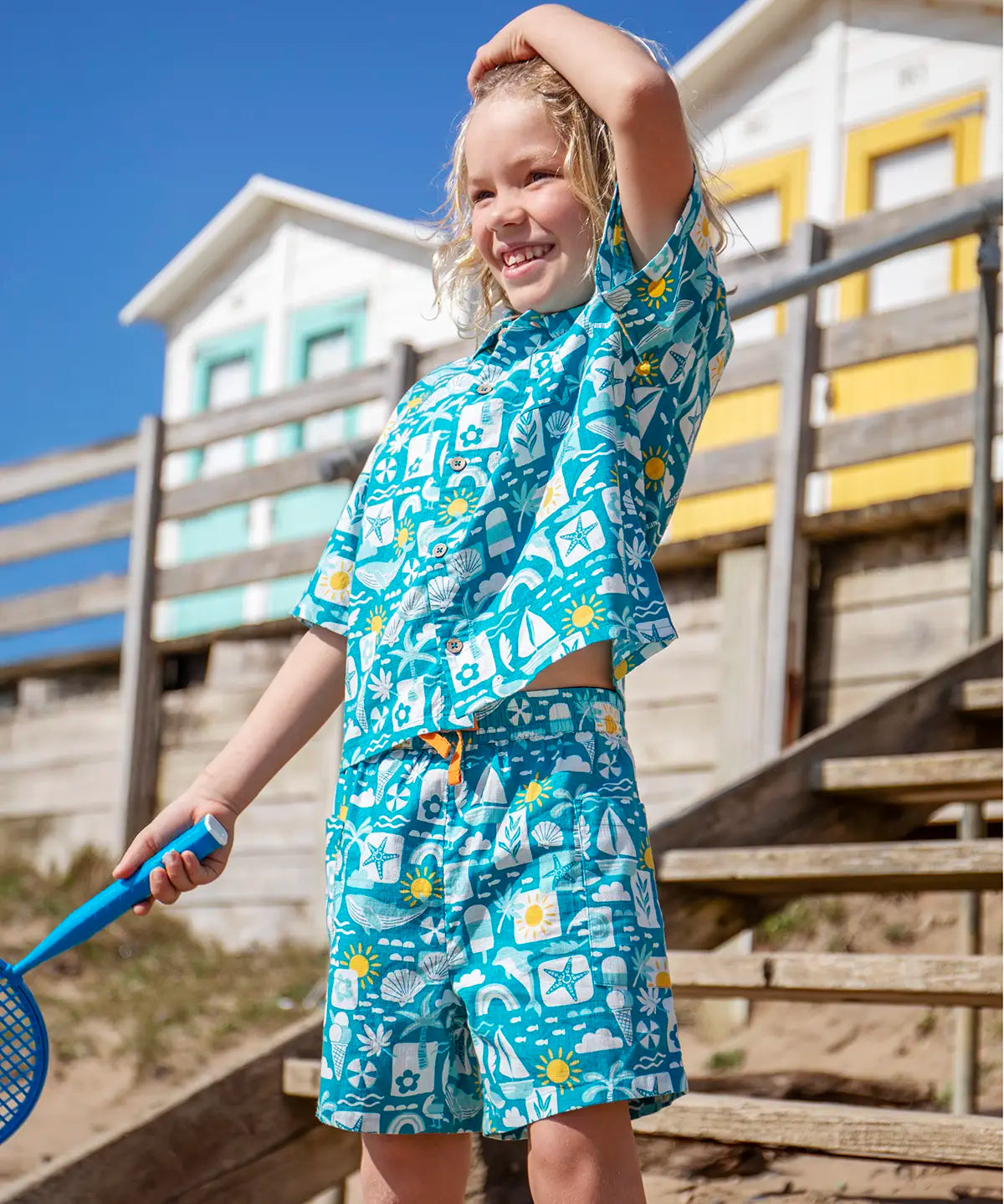 Child wearing Frugi's Kai shorts in blue with repeating beach inspired designs with beach huts in the background