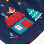 A closer look at the applique on the navy blue corduroy side of the Frugi Inara Cord Reversible Dress - Navy Blue/Rainbow Check. The house applique is blue with a red door, red roof with a Robin on top, a snowy Christmas tree. There is also embroidered holly leaf with berries, gold stars and smokey from the chimney top