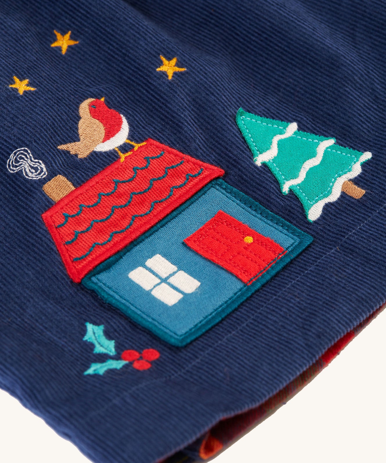 A closer look at the applique on the navy blue corduroy side of the Frugi Inara Cord Reversible Dress - Navy Blue/Rainbow Check. The house applique is blue with a red door, red roof with a Robin on top, a snowy Christmas tree. There is also embroidered holly leaf with berries, gold stars and smokey from the chimney top