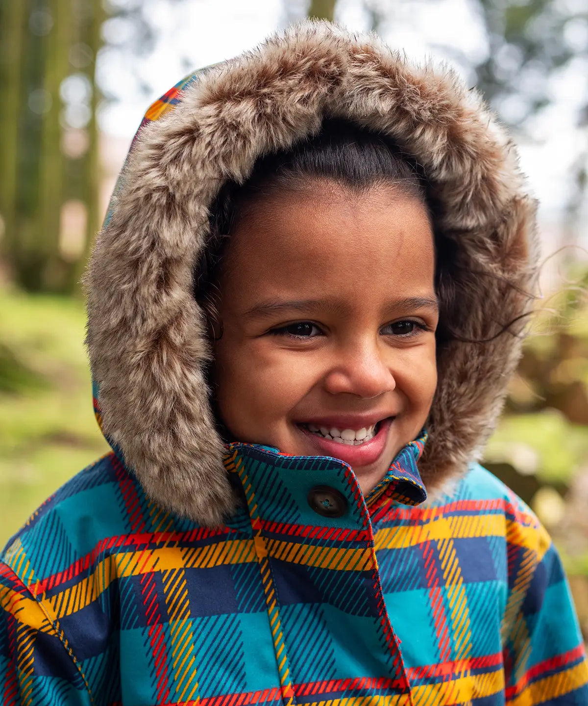 Small child wearing Frugi's Penhale parka coat in a blue red and yellow tartan check with the hood up with fur trim