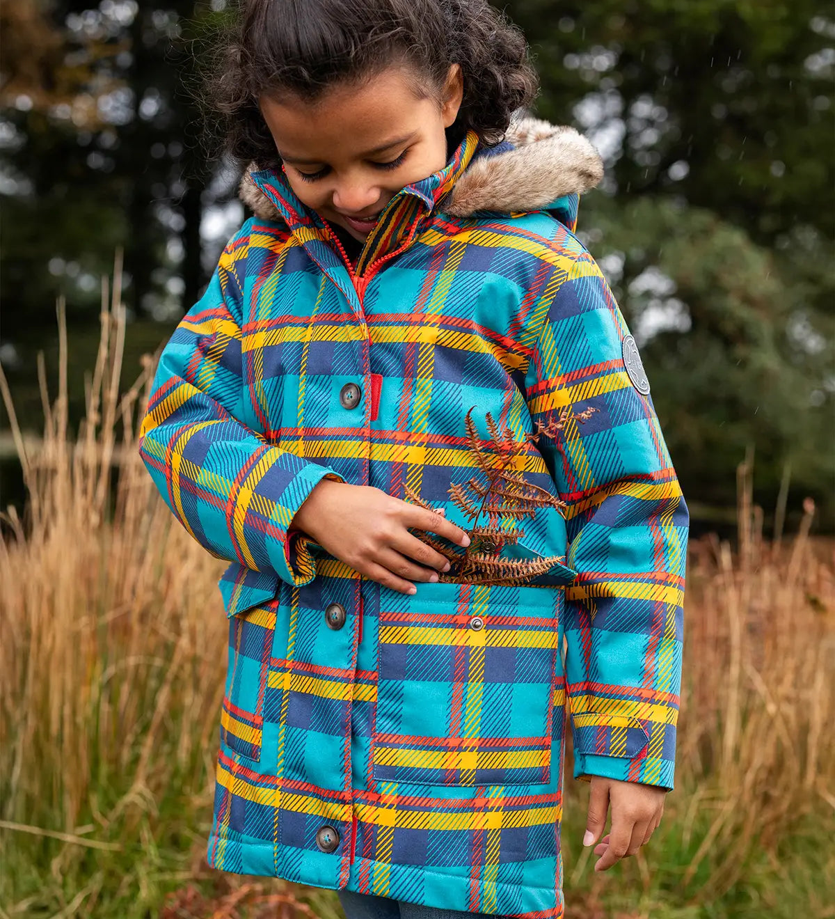 Small child wearing Frugi's Penhale parka coat in a blue red and yellow tartan check with the hood down in a grassy field