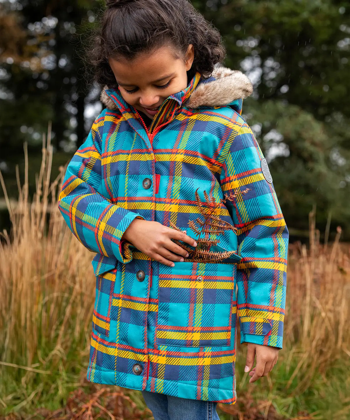 Small child wearing Frugi's Penhale parka coat in a blue red and yellow tartan check with the hood down in a grassy field