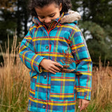 Small child wearing Frugi's Penhale parka coat in a blue red and yellow tartan check with the hood down in a grassy field