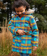 Small child wearing Frugi's Penhale parka coat in a blue red and yellow tartan check with the hood down in a grassy field