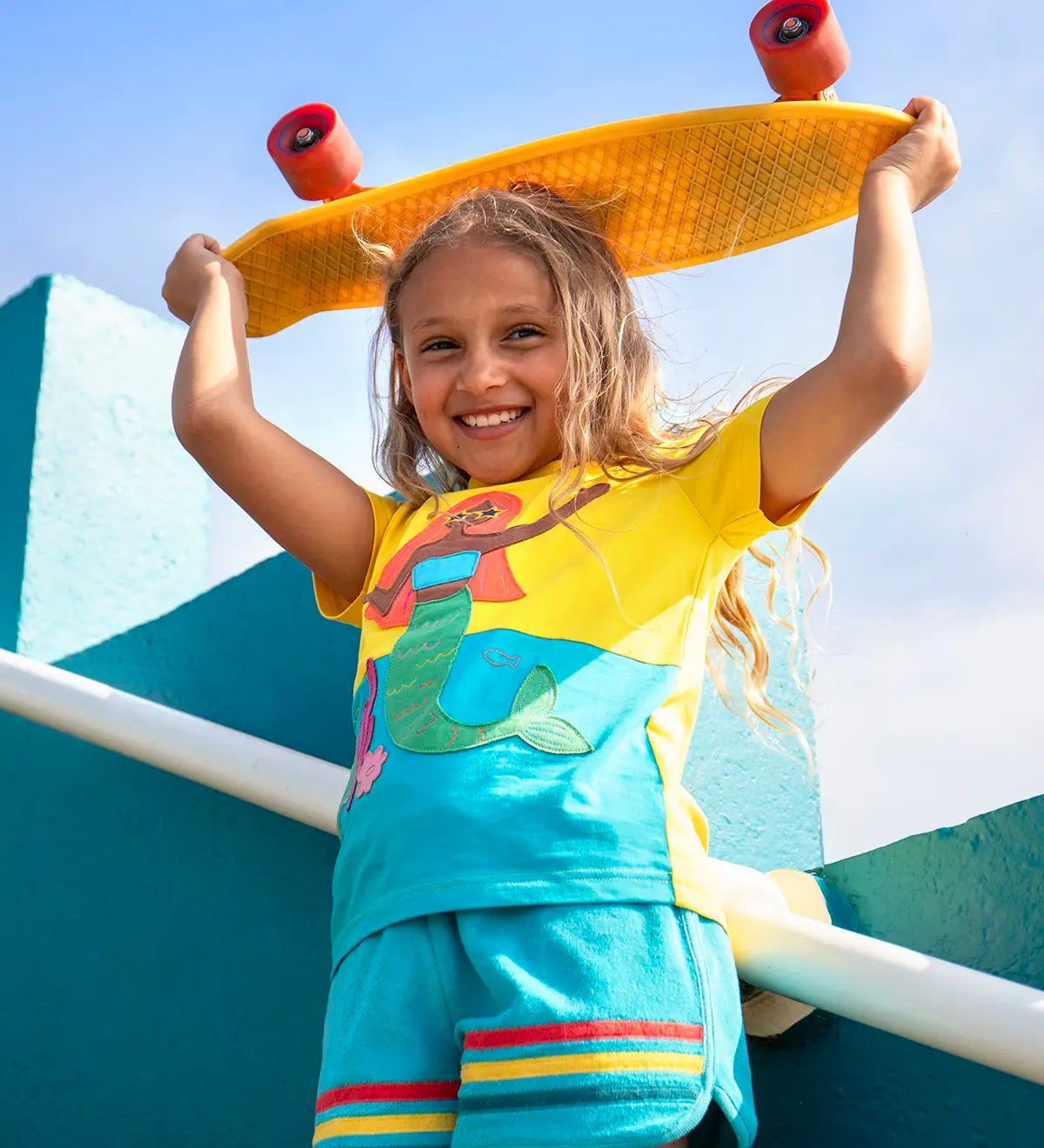 Child wearing Frugi's Poldhu hotchpotch short sleeve t-shirt in yellow and aqua with a mermaid patch and holding a skateboard