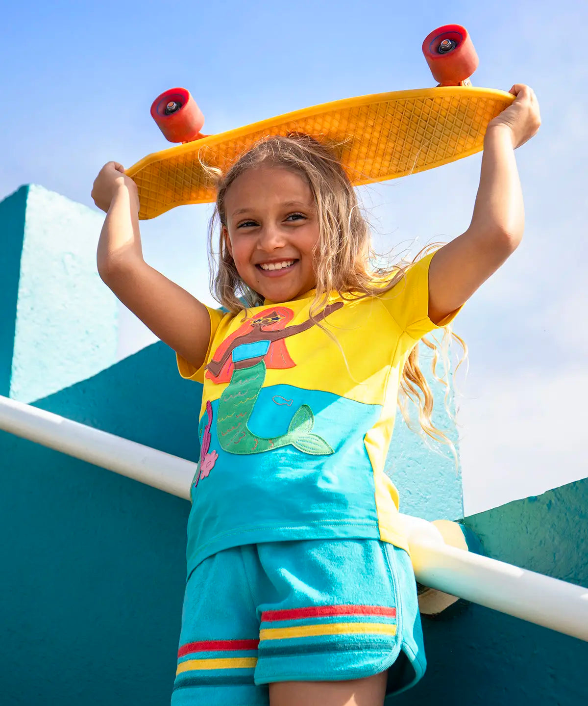 Child wearing Frugi's Poldhu hotchpotch short sleeve t-shirt in yellow and aqua with a mermaid patch and holding a skateboard