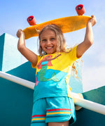 Child wearing Frugi's Poldhu hotchpotch short sleeve t-shirt in yellow and aqua with a mermaid patch and holding a skateboard