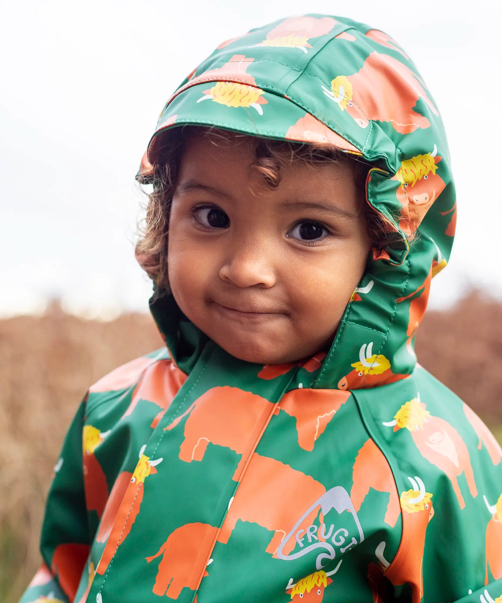 Small child wearing the Frugi waterproof green highland cow puddle buster all in one showing close up of fitted hood with extended brim