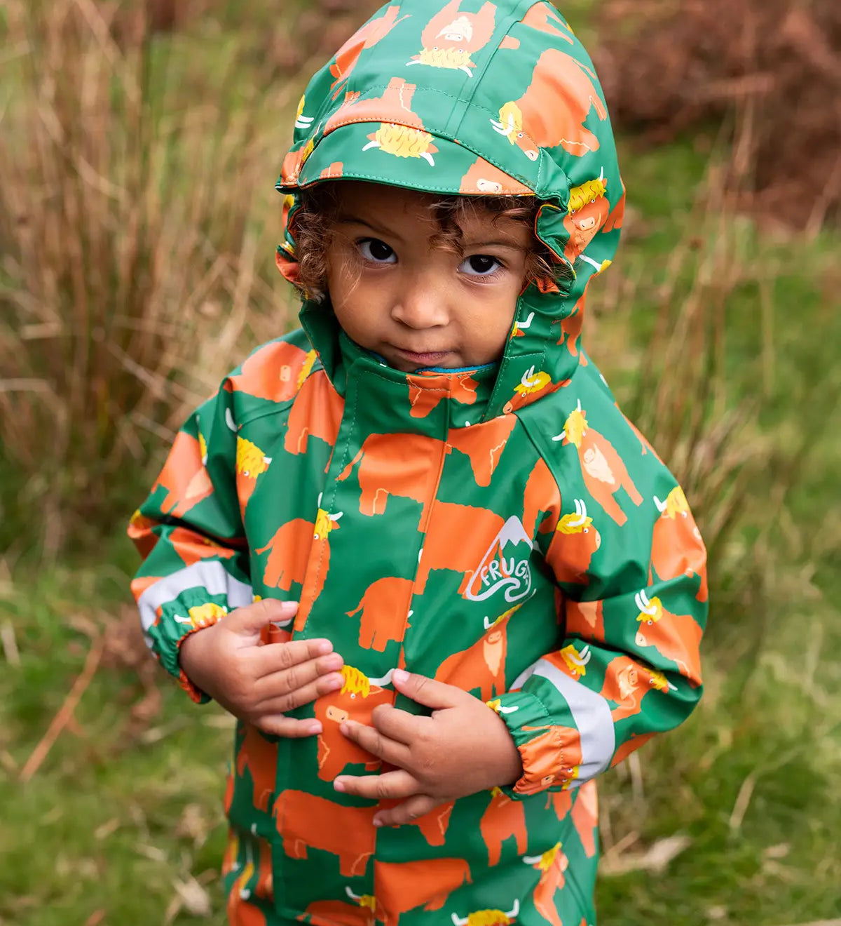 Small child wearing the Frugi waterproof green highland cow puddle buster all in one with the hood up