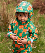 Small child wearing the Frugi waterproof green highland cow puddle buster all in one with the hood up