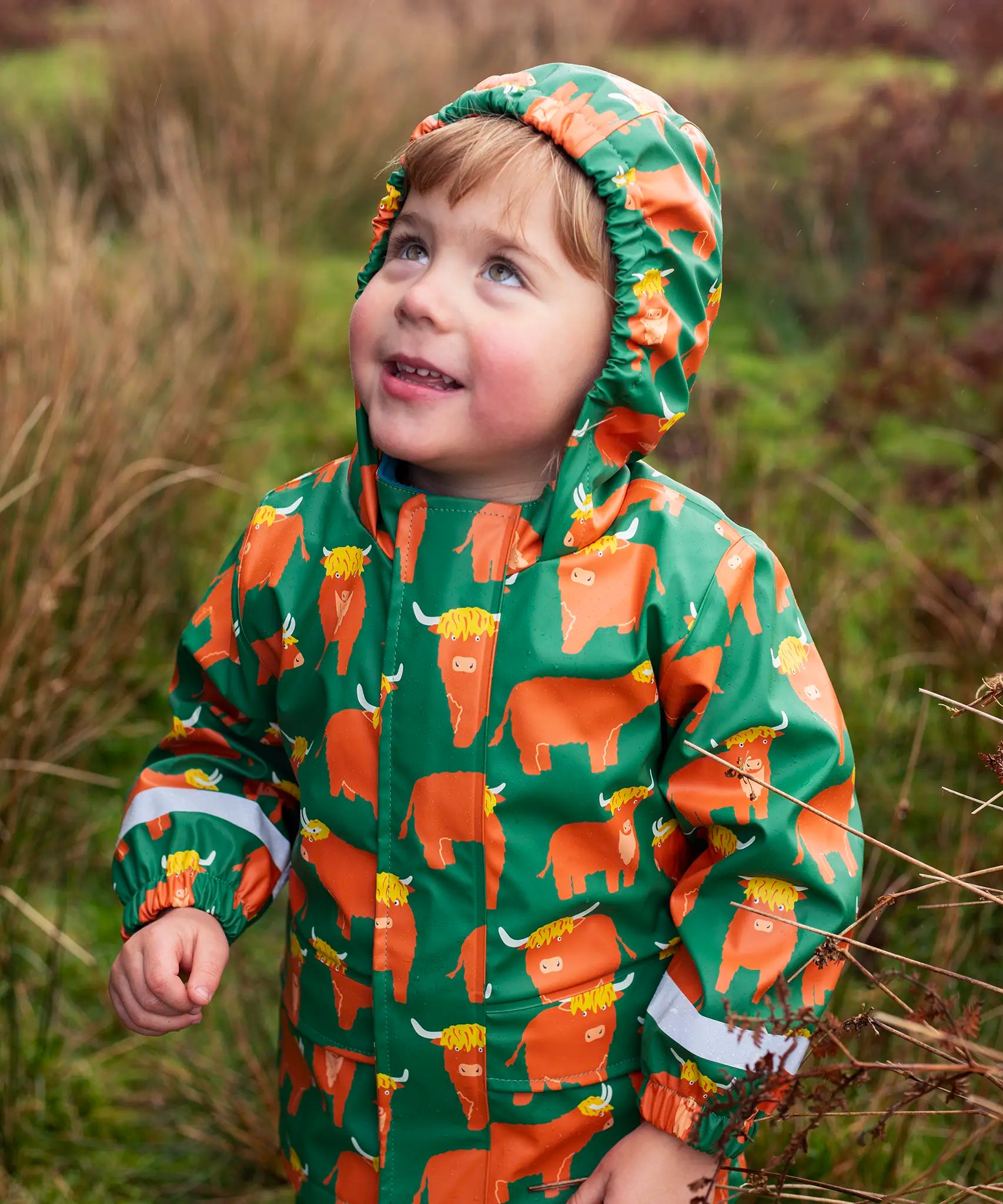 Small child wearing Frugi's waterproof puddle buster coat in highland grazing showing the fitted hood up