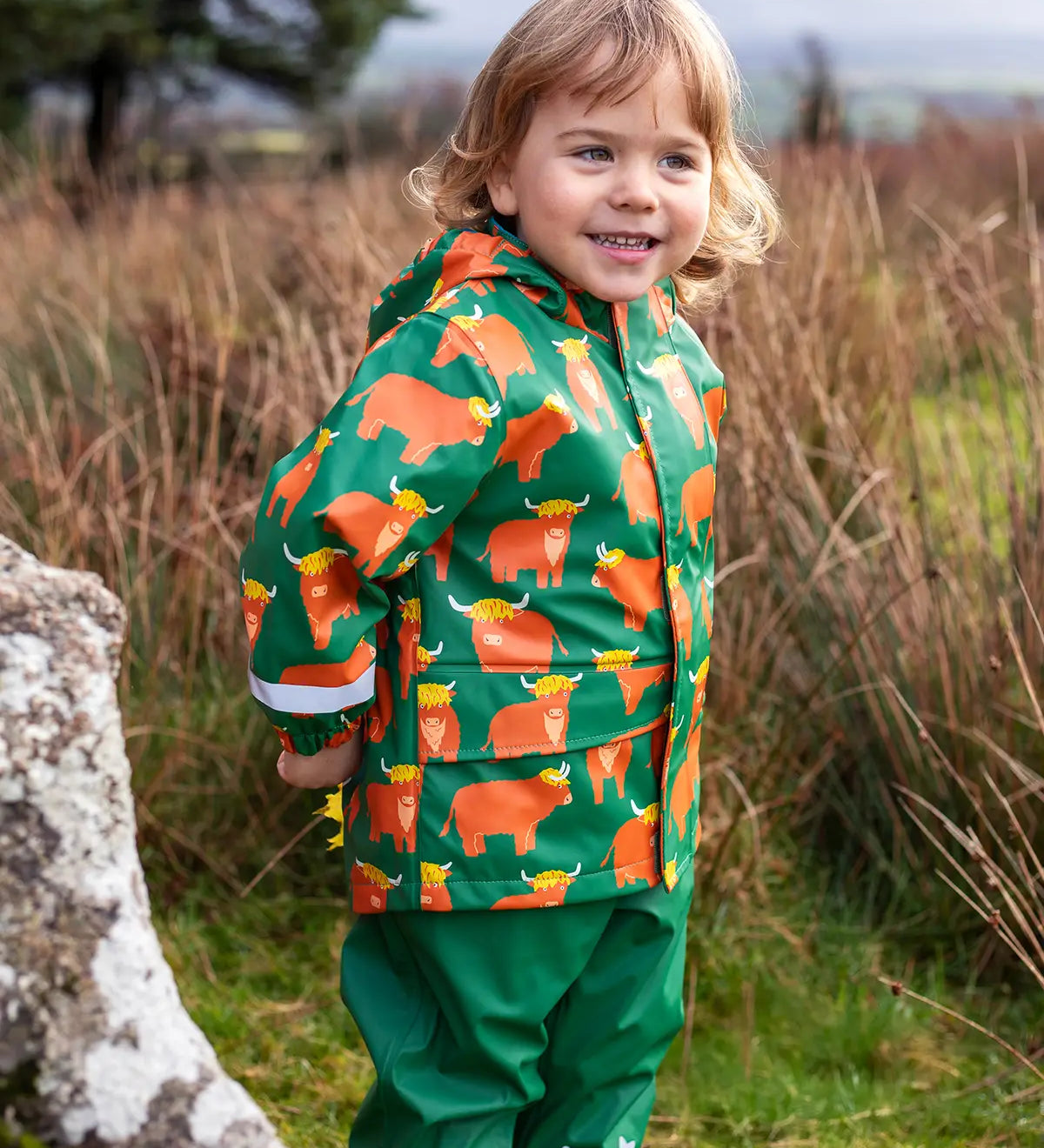 Small child wearing Frugi's waterproof puddle buster coat in highland grazing showing the side pocket and reflective strip on the cuff