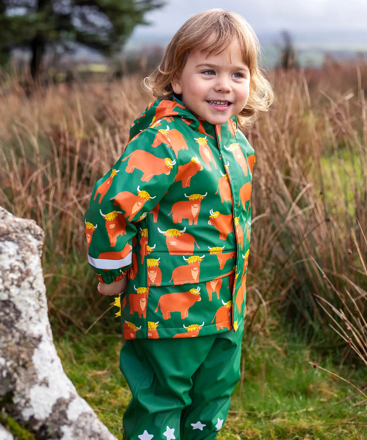Small child wearing Frugi's waterproof puddle buster coat in highland grazing showing the side pocket and reflective strip on the cuff