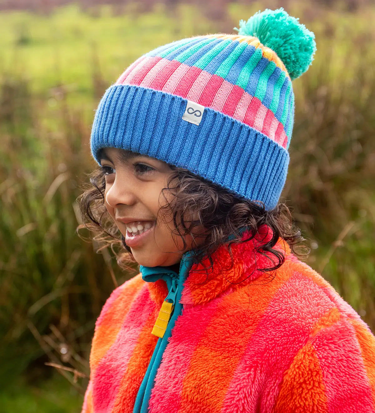 Child wearing the Frugi pastel rainbow striped bobble hat in a grassy field