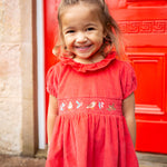A child stood in front of a red door, wearing the Frugi Amilie Party Dress - True Red/Robin