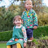 A child standing on a tree stump and wearing the Frugi snuggle fleece outfit in fun guys. A green thick fleece with yellow trim and a playful woodland mushroom print design.