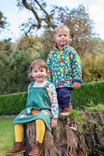 A child standing on a tree stump and wearing the Frugi snuggle fleece outfit in fun guys. A green thick fleece with yellow trim and a playful woodland mushroom print design.