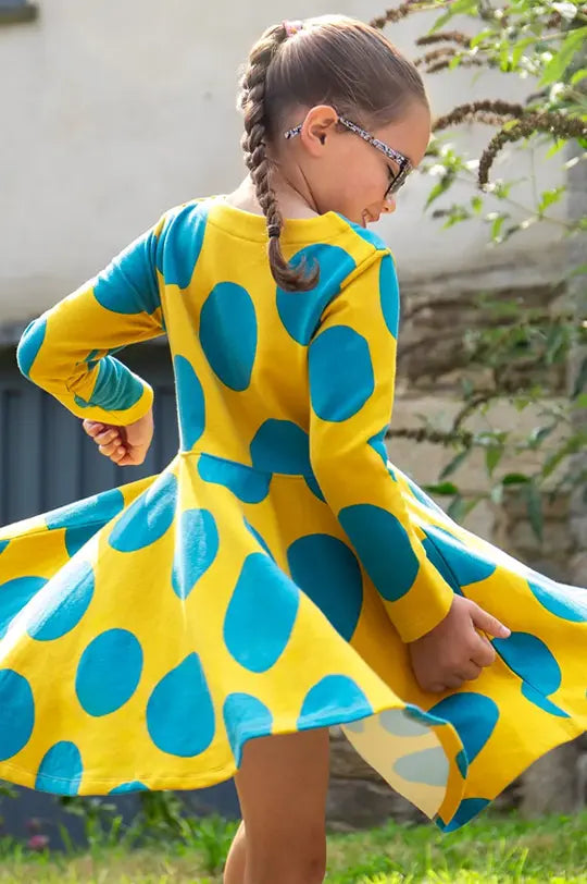 Sale on Frugi Kids clothing at Babipur. Represented with Frugi Organic yellow dress with blue polka dots, child standing outdoors.
