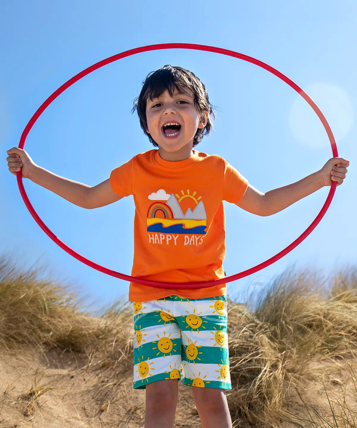 Child wearing Frugi's green and white striped shorts with repeating sun design  and a orange happy days applique tshirt