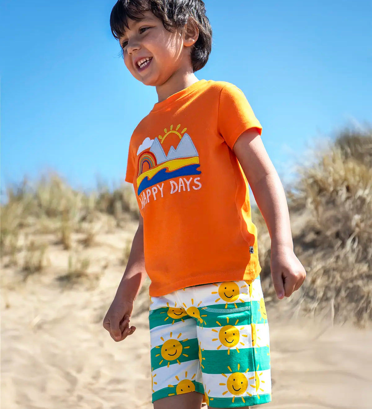 Child wearing Frugi's green and white striped shorts with repeating sun design  and a orange happy days applique tshirt