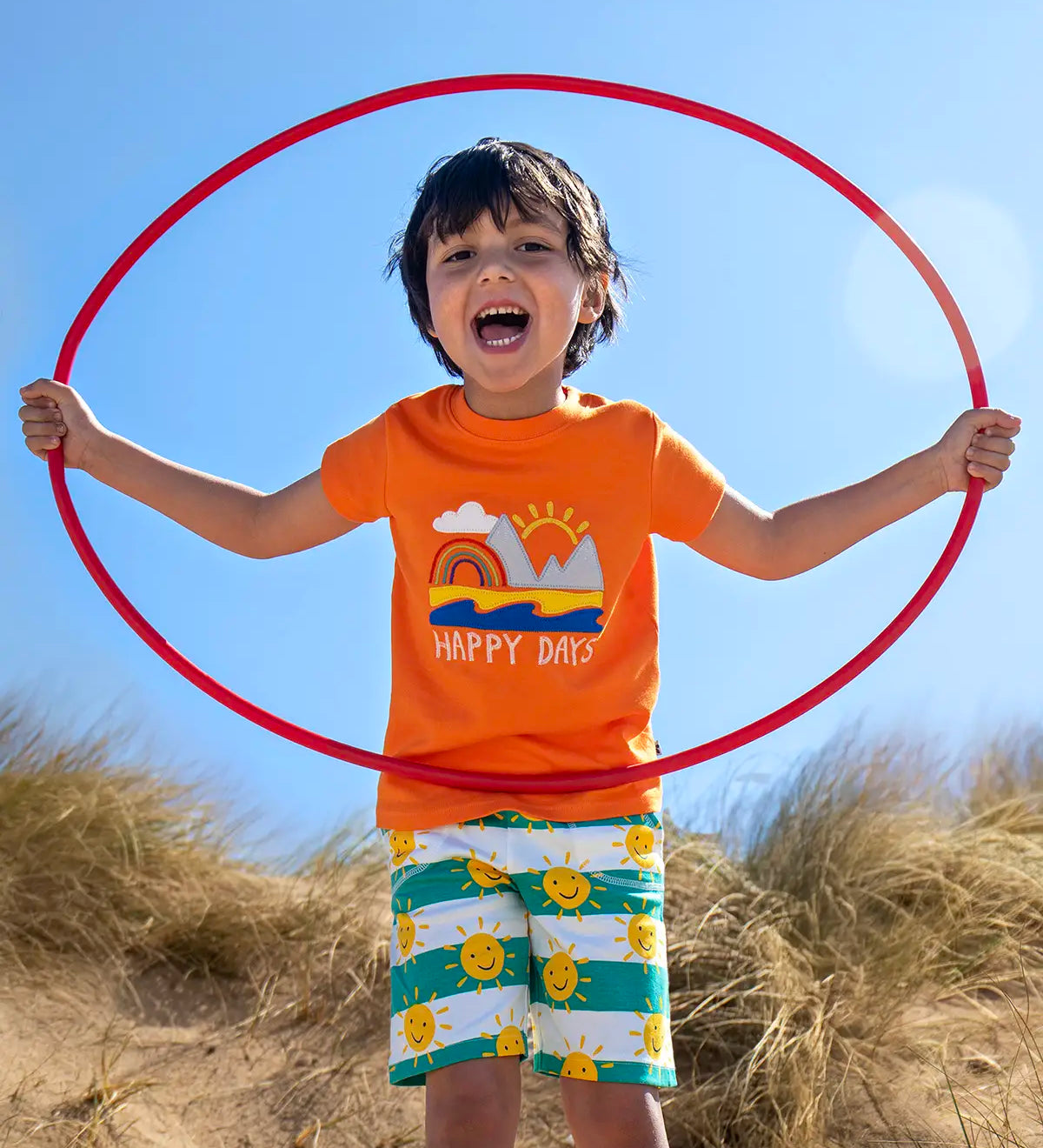 Child wearing Frugi's green and white striped shorts with repeating sunshine design on the beach holding a red hoop