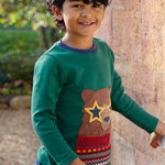 A child, stood outside next to a large brick wall, wearing the Frugi Terrific Applique Top - Teal/Fairisle Bear