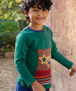 A child, stood outside next to a large brick wall, wearing the Frugi Terrific Applique Top - Teal/Fairisle Bear