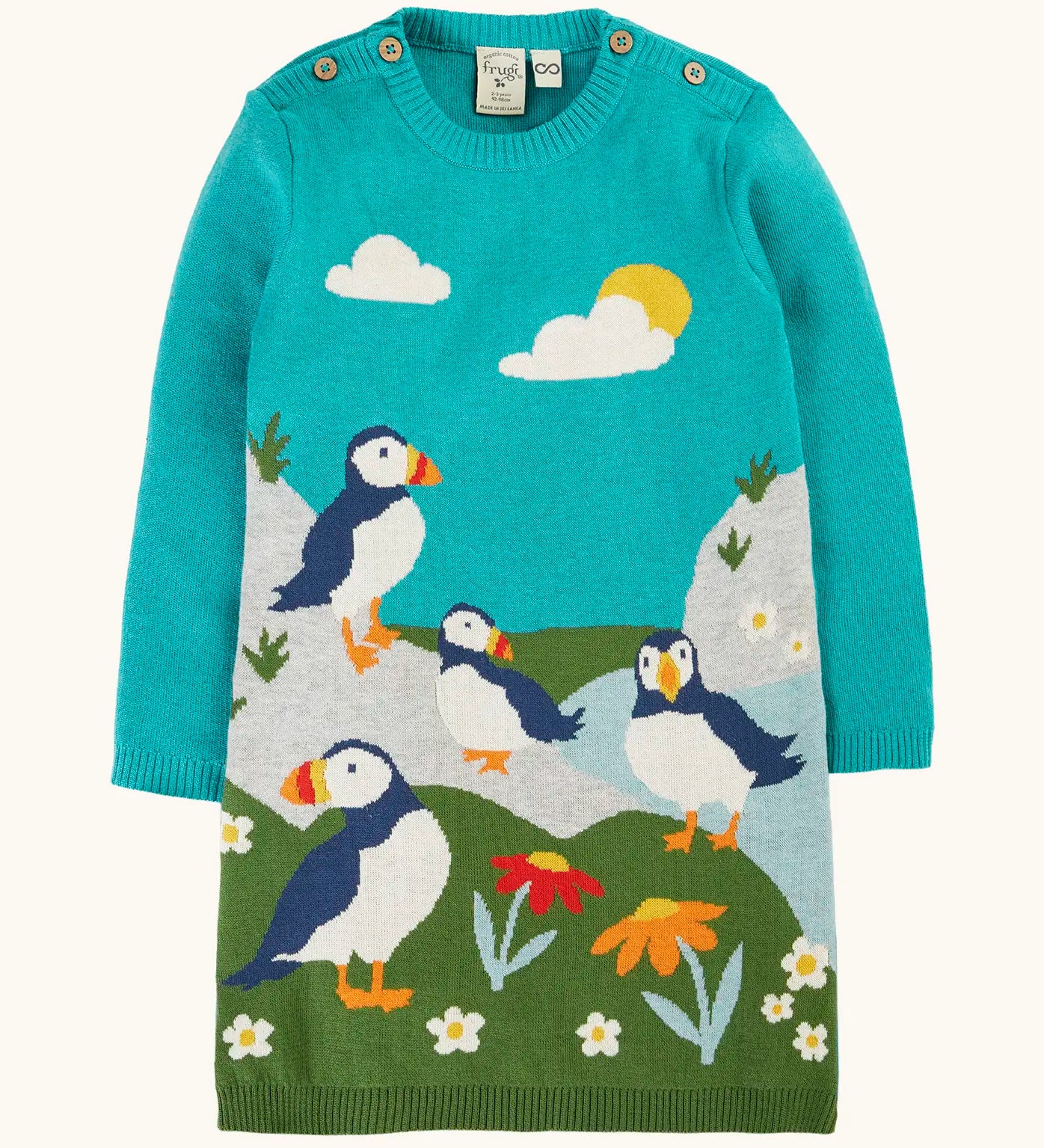 Frugi GOTS organic cotton kids dress in light blue with puffins standing on grass with flowers . Features button fasteners on the shoulders