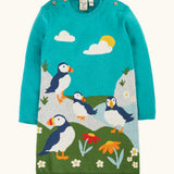 Frugi GOTS organic cotton kids dress in light blue with puffins standing on grass with flowers . Features button fasteners on the shoulders