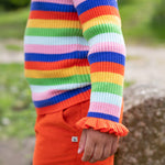 A child is stood outside, wearing the Frugi Zoe Knitted Jumper - Retro Stripe. The image shows a close up of the child's arm and the ruffled wrist cuffs cover over the top of the hand. The colour-block stripes look very vibrant n the natural day light