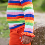 A child is stood outside, wearing the Frugi Zoe Knitted Jumper - Retro Stripe. The image shows a close up of the child's arm and the ruffled wrist cuffs cover over the top of the hand. The colour-block stripes look very vibrant n the natural day light