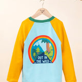 The back of the Frugi Children's Leo raglan long sleeved top hanging on a branch and available at Babipur.. The body of the top is light blue with yellow gold coloured sleeves and a contrasting dark green neck collar. The large circular graphic on the back features the words out in the wild written on a river surrounded by land, trees, mountains all surrounded by a three colour rainbow.