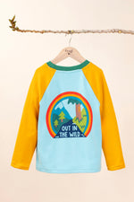 The back of the Frugi Children's Leo raglan long sleeved top hanging on a branch and available at Babipur.. The body of the top is light blue with yellow gold coloured sleeves and a contrasting dark green neck collar. The large circular graphic on the back features the words out in the wild written on a river surrounded by land, trees, mountains all surrounded by a three colour rainbow.