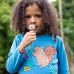 A child blowing a dandelion, whilst wearing the Frugi Adventure Applique Top - Deep Water/Squirrel