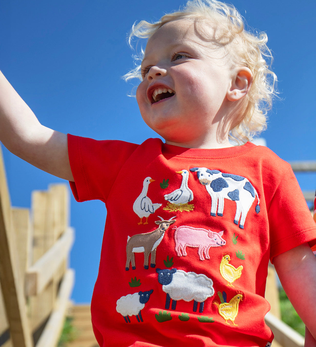 A child smiling with wooden stairs in the background. Wearing the red Frugi short sleeve t-shirt with different farm animal patches on the front.