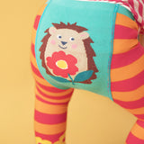 A close up of the back of the Frugi orange and pink striped knitted with a hedgehog holding a flower design worn by a child standing in front of a light orange background 