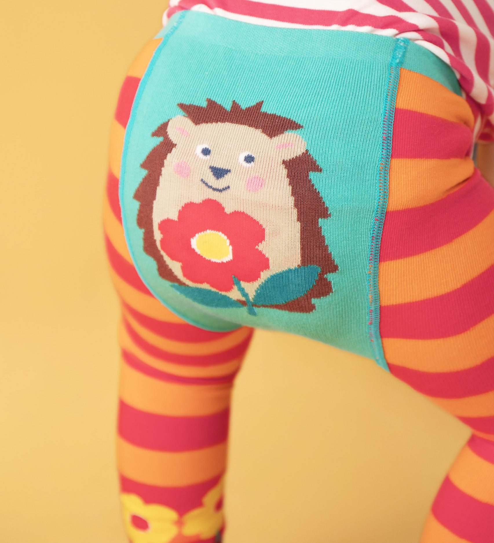 A close up of the back of the Frugi orange and pink striped knitted with a hedgehog holding a flower design worn by a child standing in front of a light orange background 