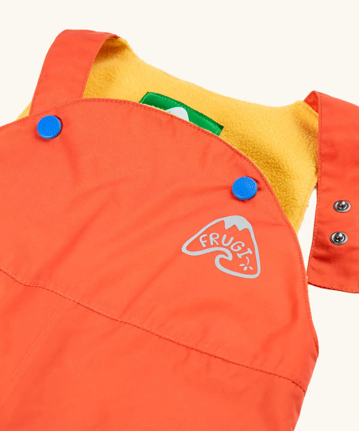 Frugi little waterproof dungarees in fire orange laid flat showing open popper fastener on the shoulder strap for size adjustment