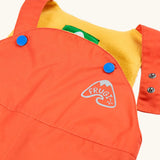 Frugi little waterproof dungarees in fire orange laid flat showing open popper fastener on the shoulder strap for size adjustment