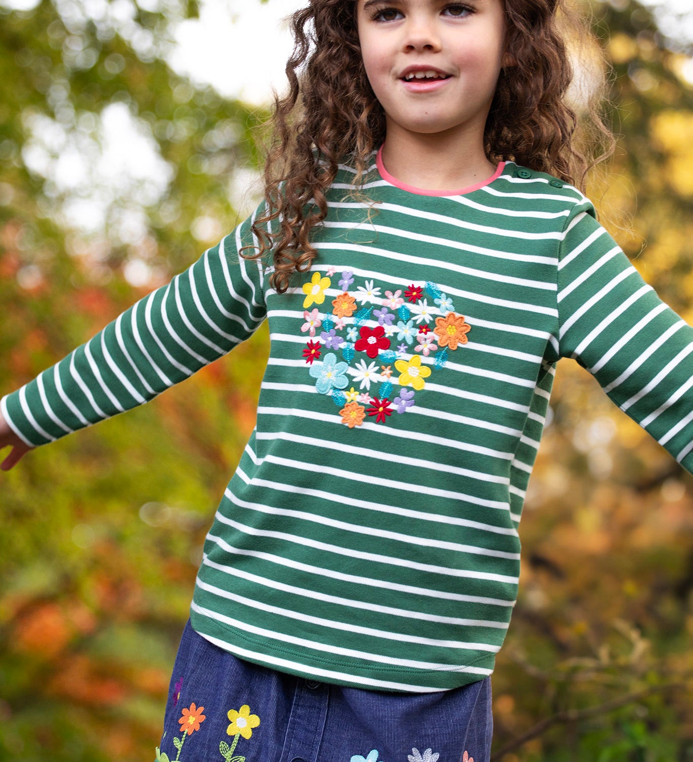 A child standing outside and wearing the Frugi Louise embroidered top in green breton / heart. A GOTS organic cotton top available at Babipur in green and white stripe with a collection of embroidered flowers shaped as a heart