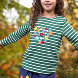 A child standing outside and wearing the Frugi Louise embroidered top in green breton / heart. A GOTS organic cotton top available at Babipur in green and white stripe with a collection of embroidered flowers shaped as a heart