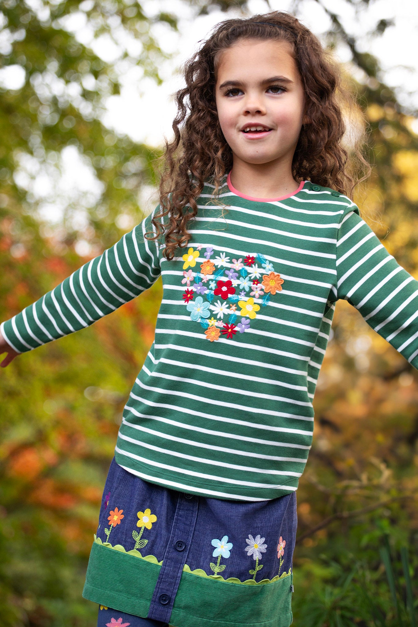 A child standing outside and wearing the Frugi Louise embroidered top in green breton / heart. A GOTS organic cotton top available at Babipur in green and white stripe with a collection of embroidered flowers shaped as a heart