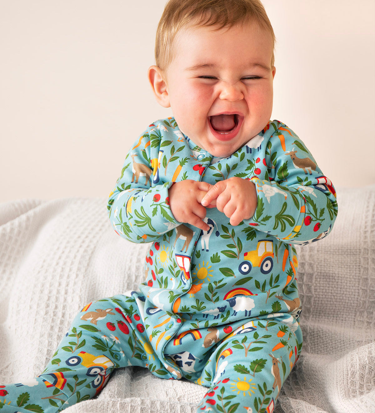 A baby laughing and sitting on a soft grey blanket. Wearing the light blue frugi footed babygrow with a repeating farm themed print