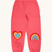 Frugi organic cotton joggers in pink with a heart patch on one knee and a rainbow on the other. Shows 2 front pockets