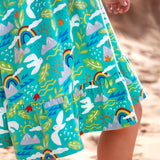 Child standing on sand wearing Frugi's skater dress in aqua with a repeating seagull and mountain design showing the hem