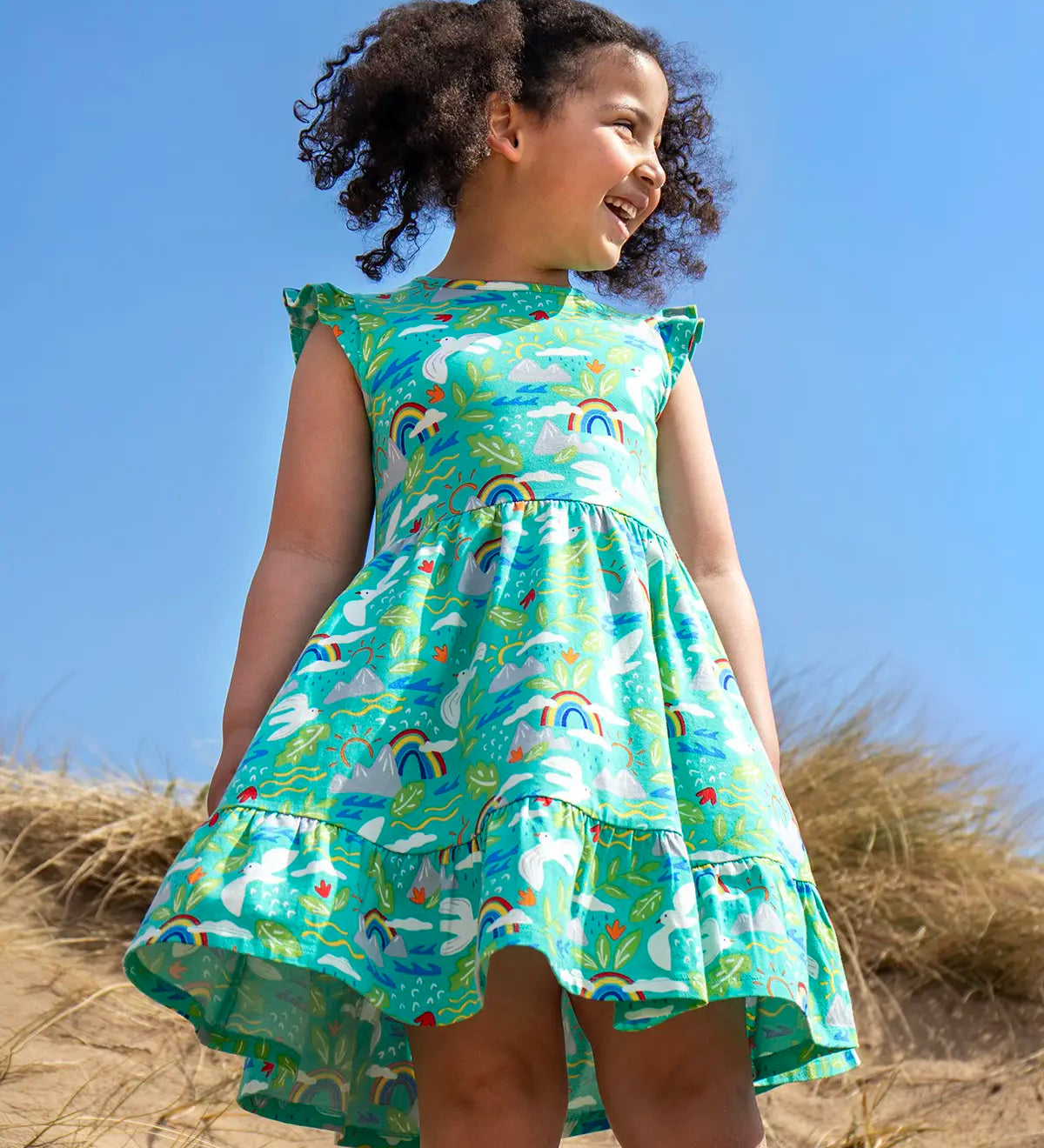 Child wearing Frugi's skater dress in aqua with a repeating seagull and mountain design on the beach 