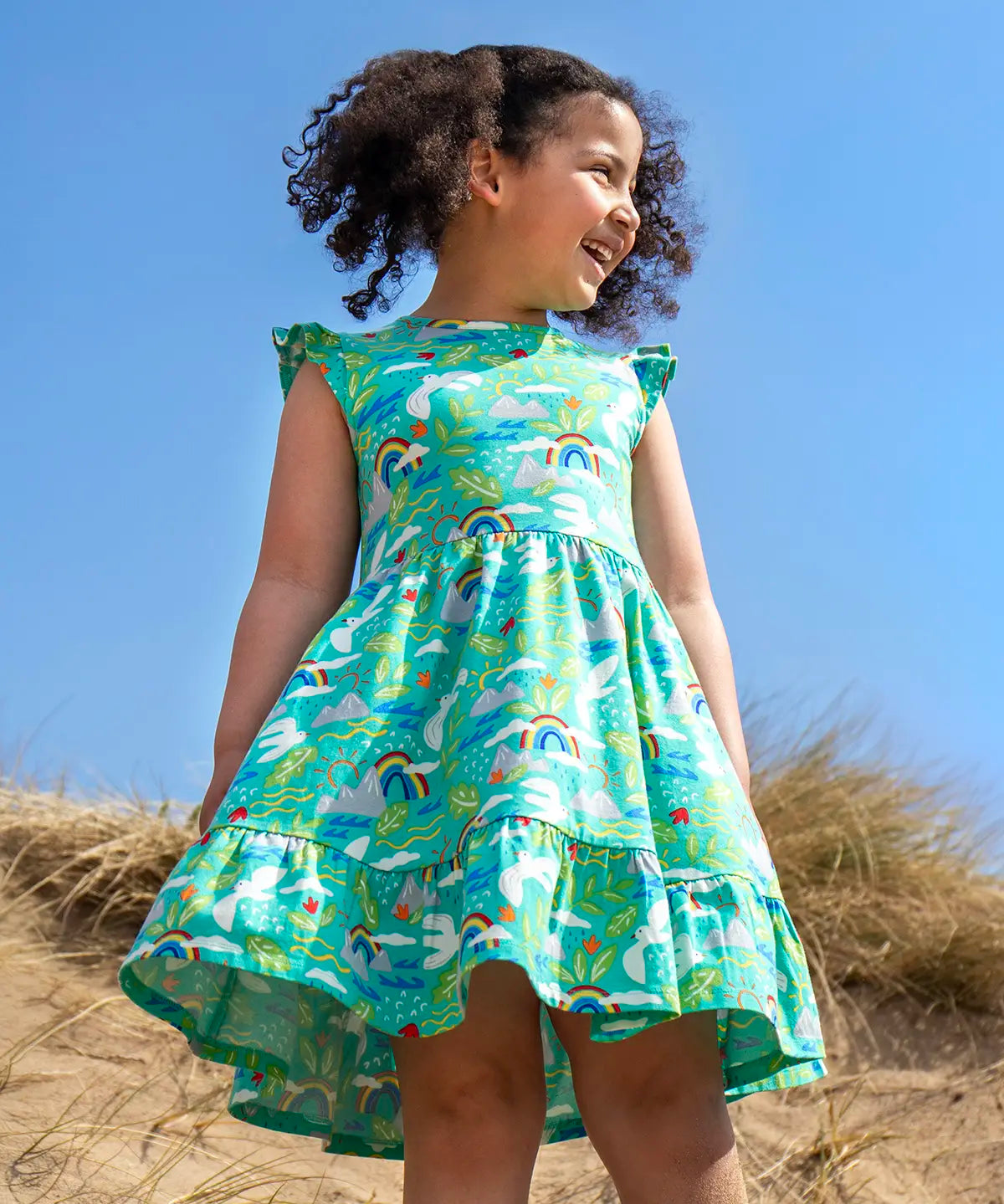 Child wearing Frugi's skater dress in aqua with a repeating seagull and mountain design on the beach 